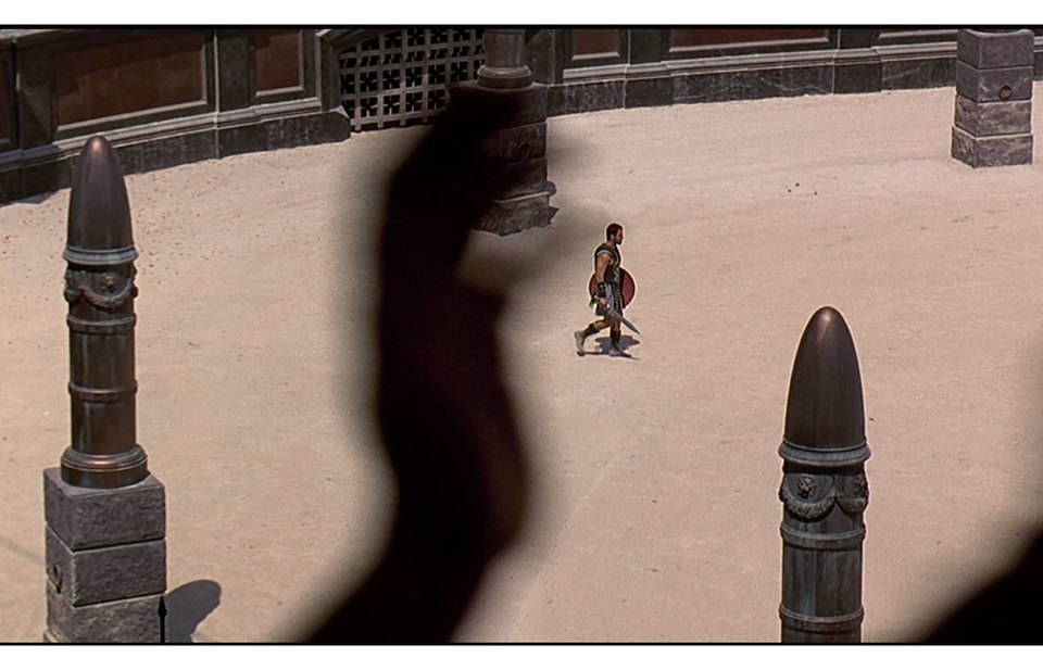 High-angle Gladiator reference frame of a lone gladiator crossing the arena sand, partly obscured by foreground architecture.